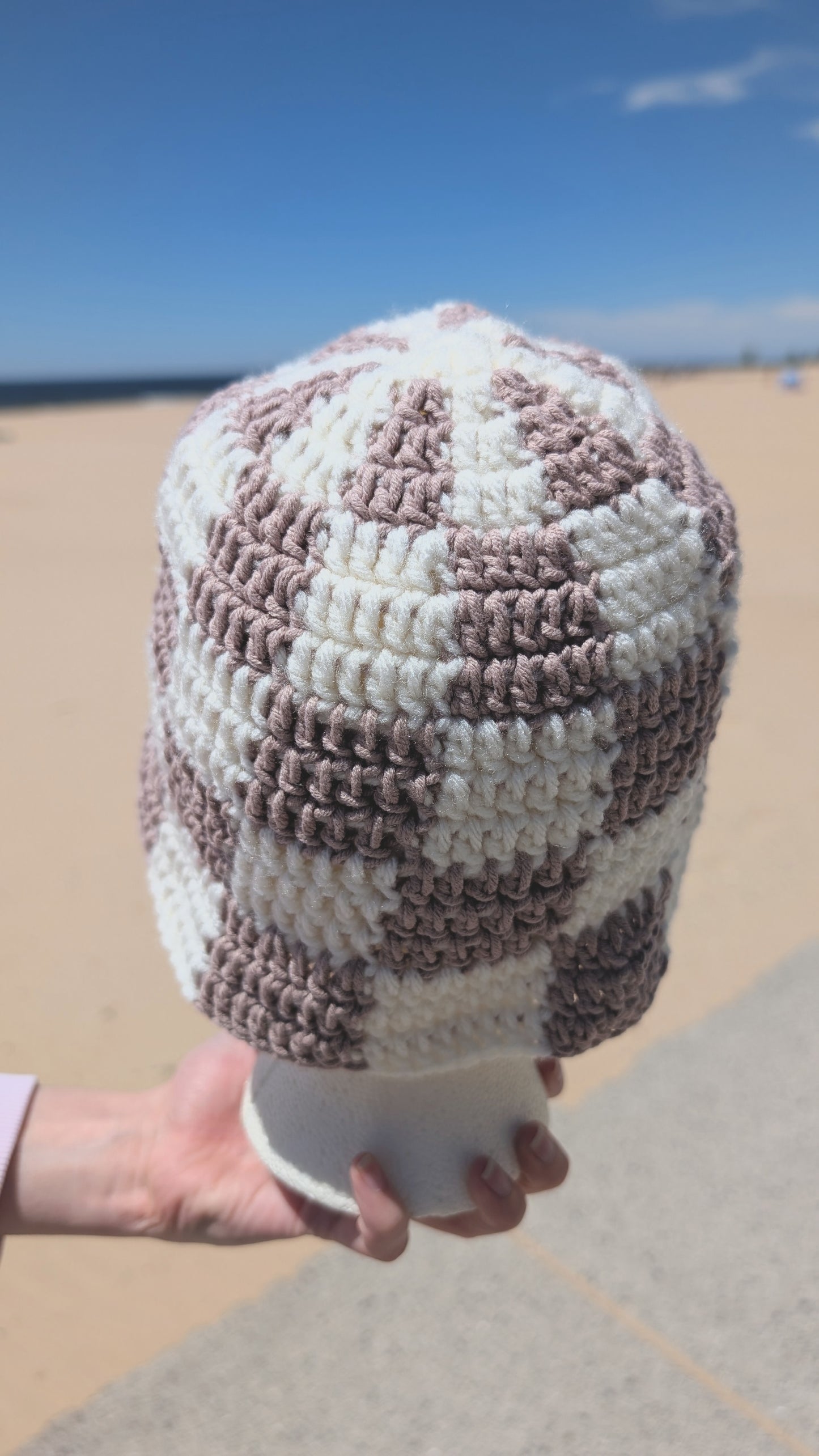 Hand holding a crocheted bucket hat with a checkered pattern on a beach.