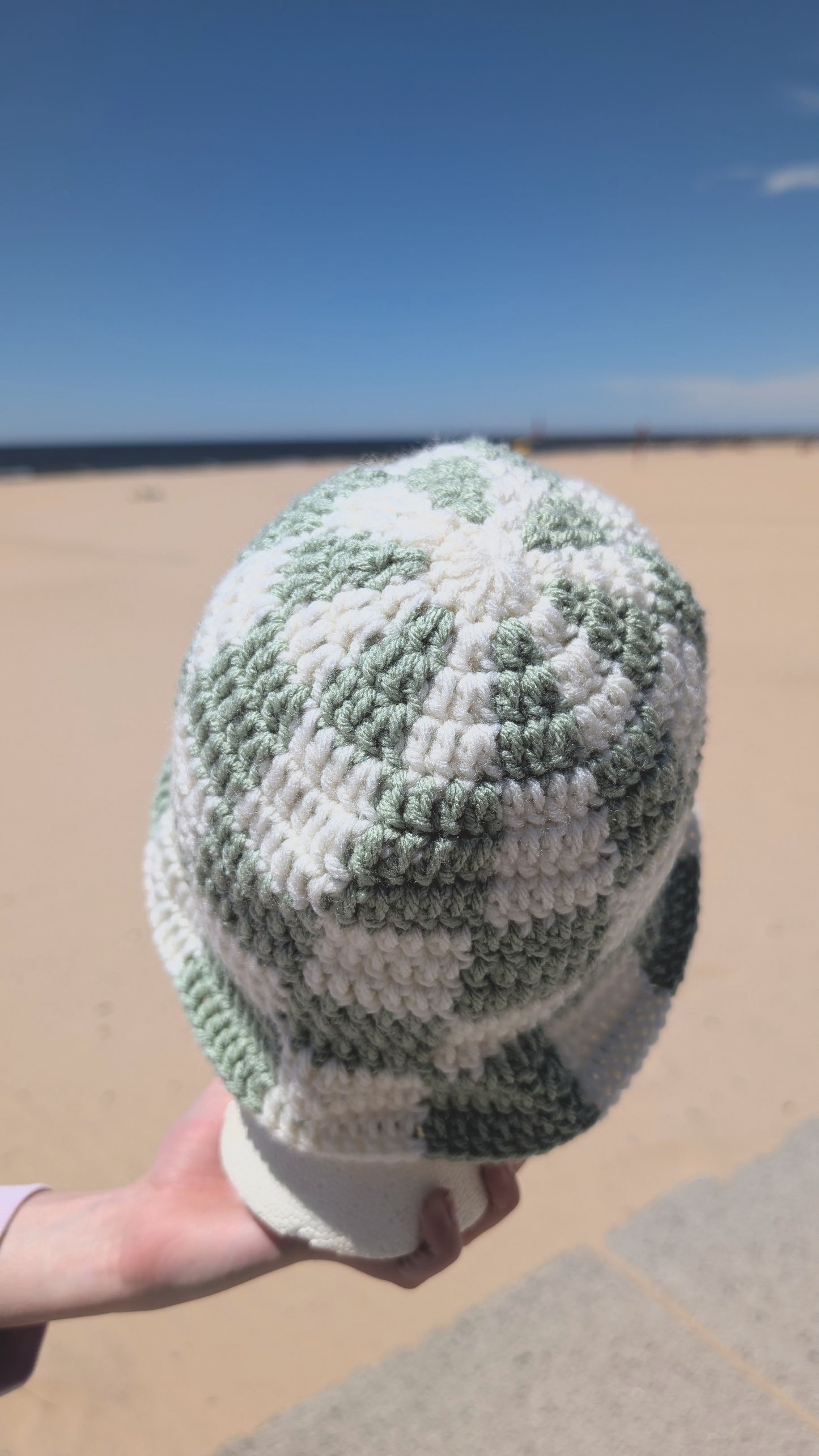 Green and white patterned beanie held by a hand on a beach.