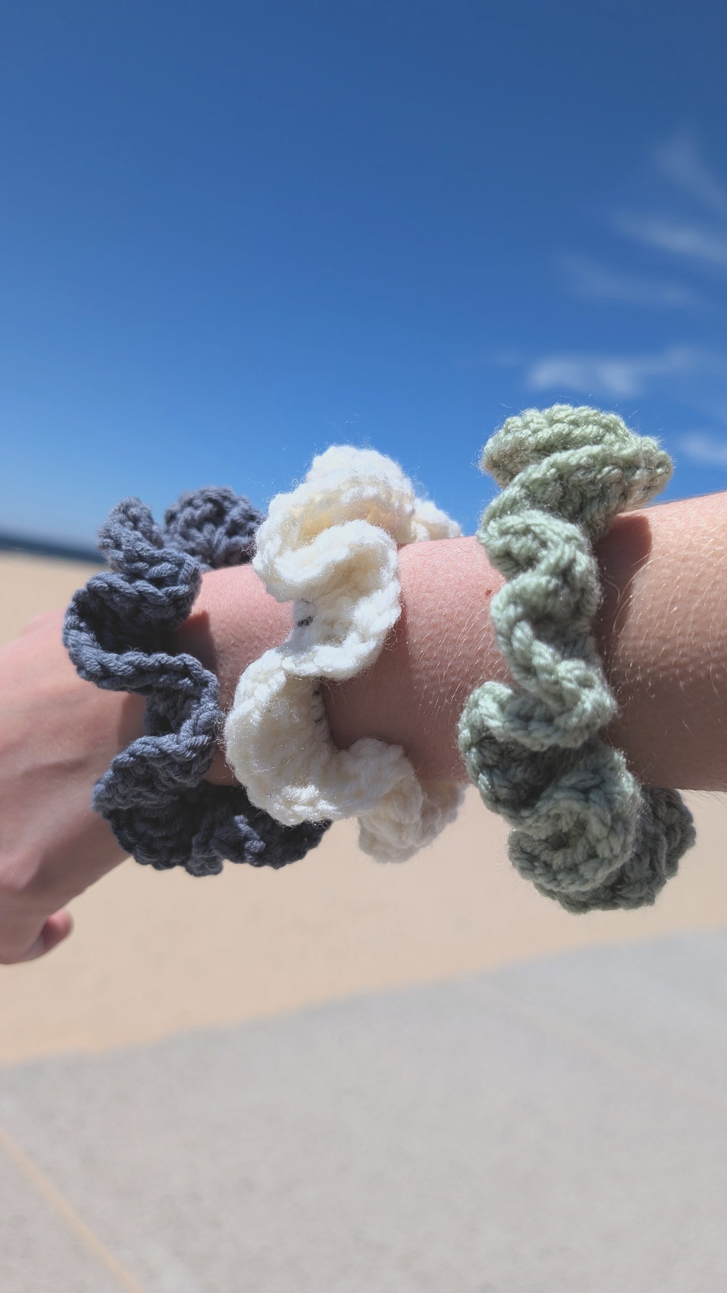Three crocheted hair ties in blue, white, and green held against a clear blue sky.
