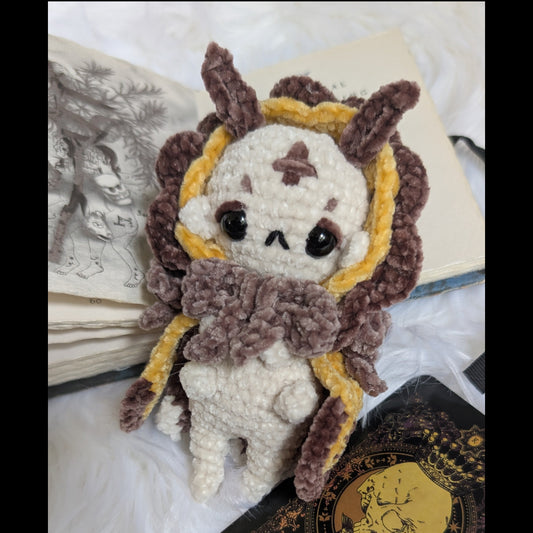 Handmade crochet moth-fairy plush in amber, caramel, and cream on a warm autumn backdrop